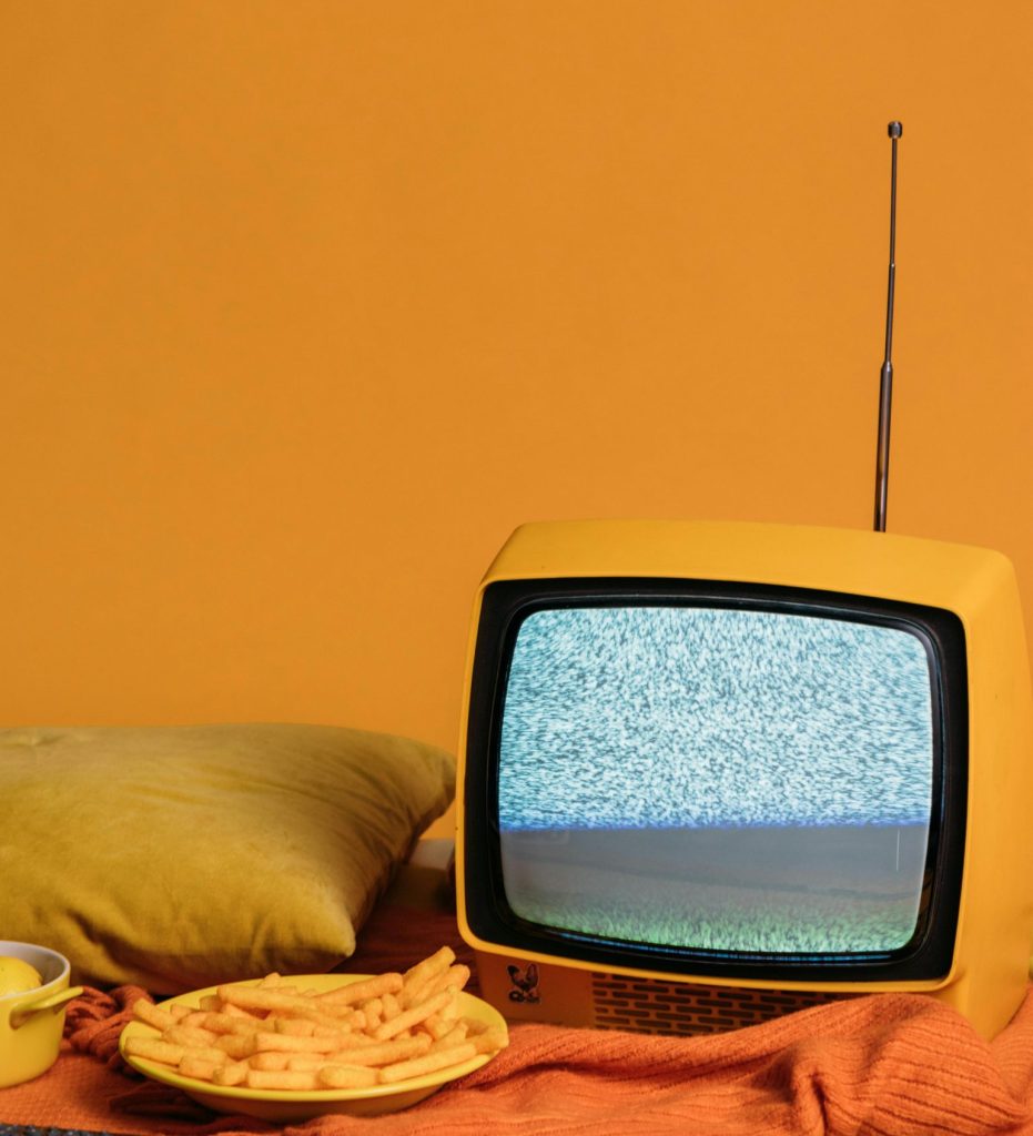 Vintage TV with static on screen surrounded by fries, lemons, and pillow on yellow backdrop.